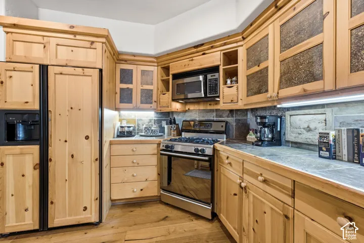 Natural wood cabinetry with integrated paneled refrigerator, stainless steel appliances, stone tile backsplash, and light-toned countertops
