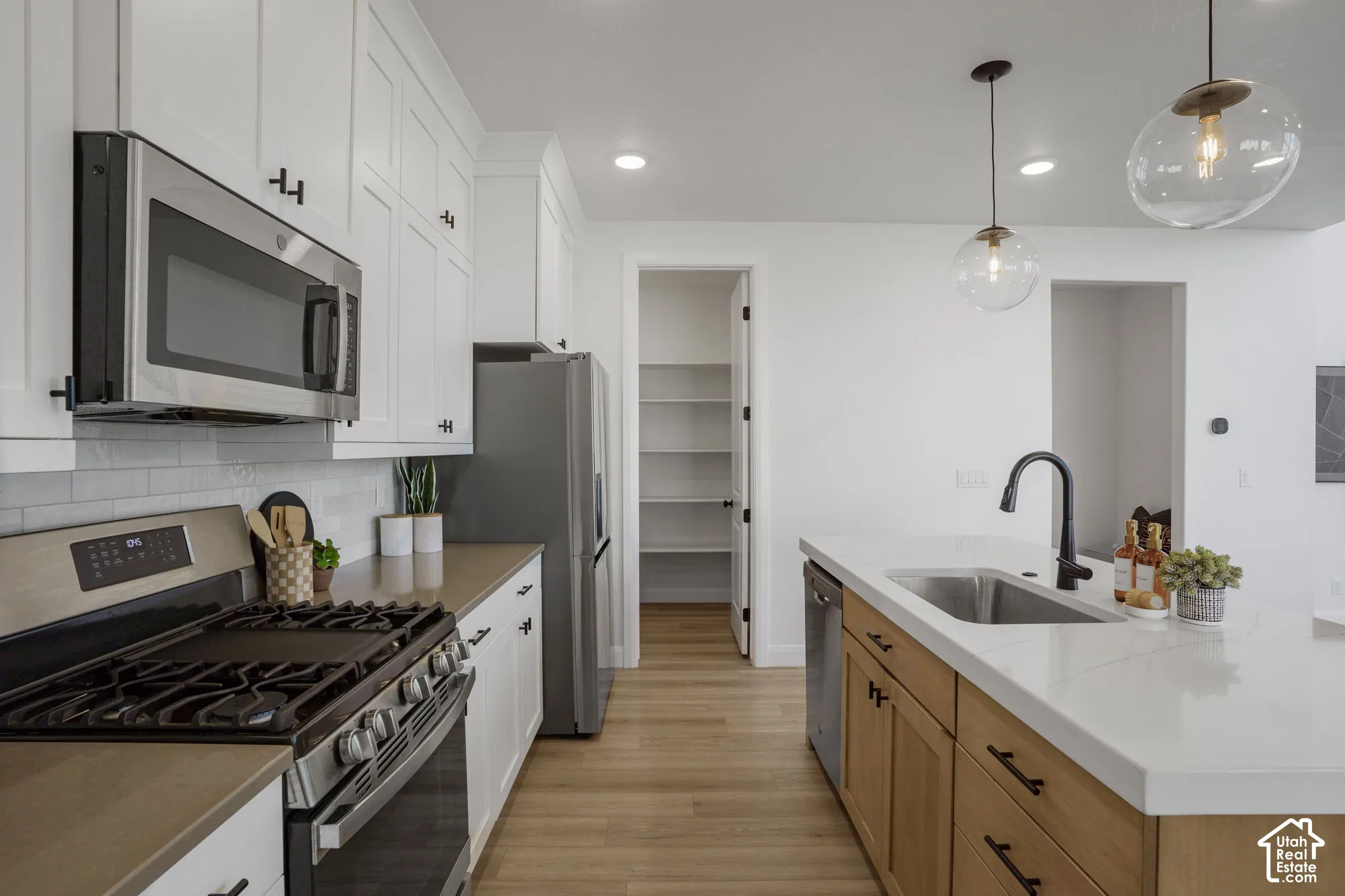 White shaker cabinetry, stainless steel appliances, wood-finish flooring, central island with sink, and a walk-in pantry