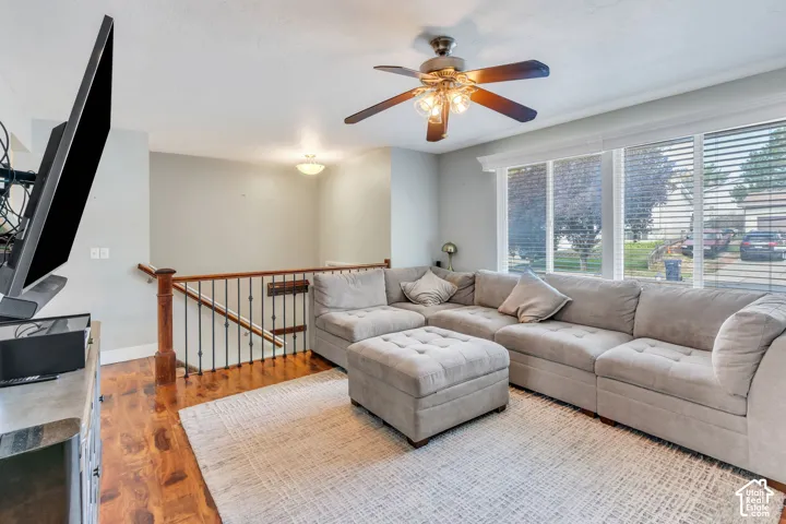 Wood-finish flooring with a light-toned area rug, a ceiling fan with integrated lighting, and large windows with blinds