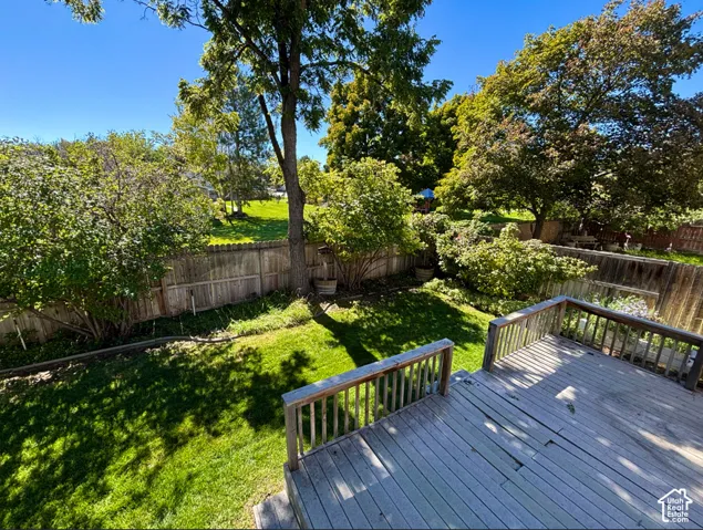 Wood deck with railing overlooking a fenced backyard featuring a manicured lawn and mature trees