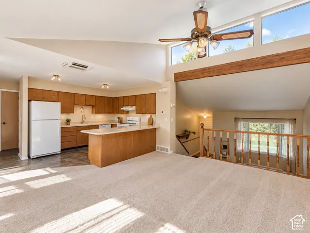 Open-concept living space featuring a kitchen with wood cabinetry and a breakfast bar, carpeted flooring, a ceiling fan, and high windows providing natural illumination