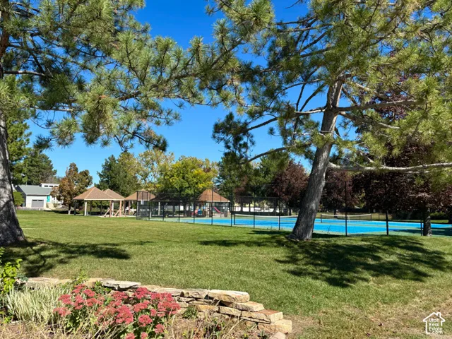 Expansive green space featuring mature trees, a stone retaining wall with plantings, and a tennis court with perimeter fencing
