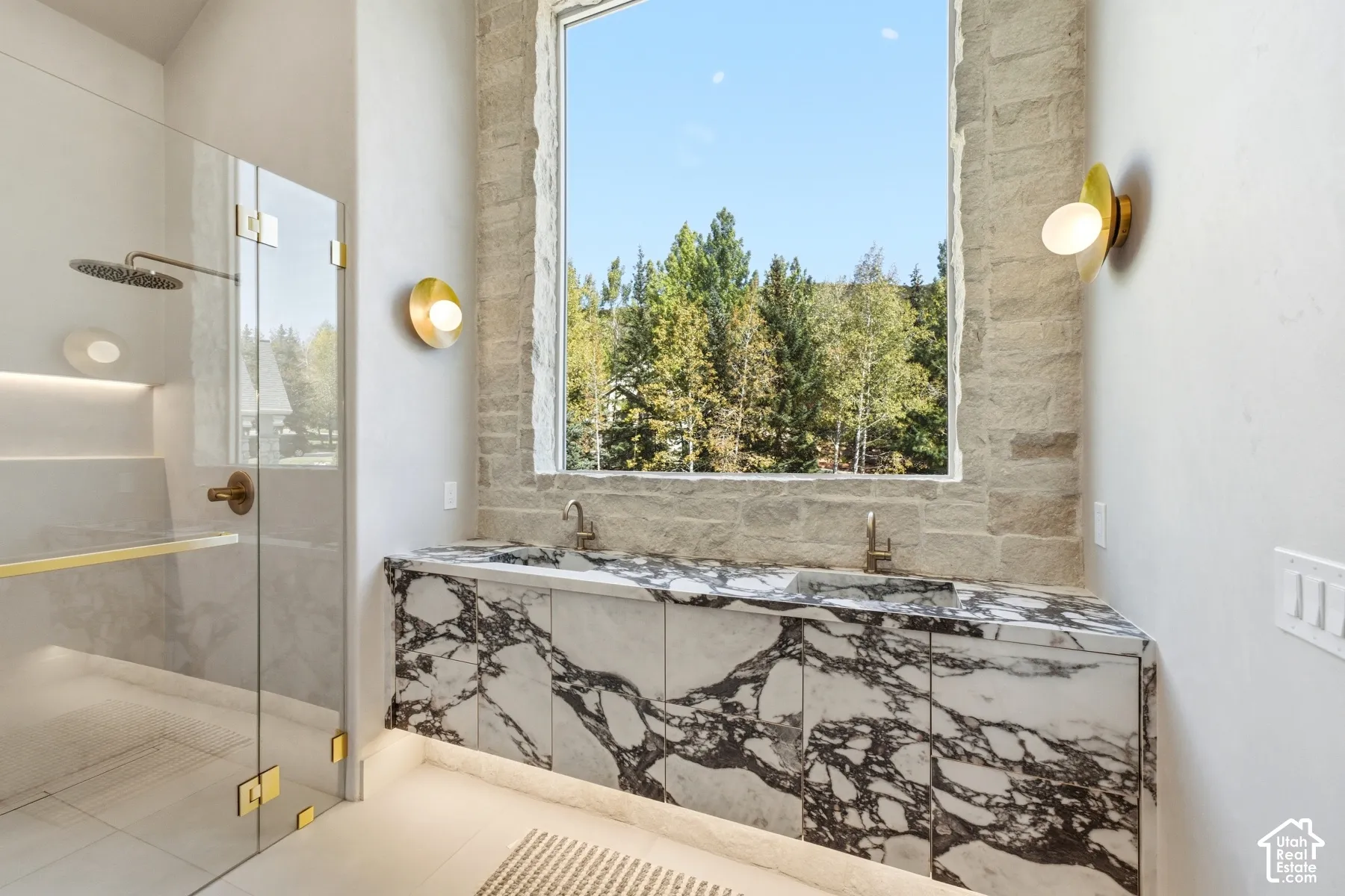 Oversized window with stone surround, dual vanity with marble countertop and integrated sinks, glass-enclosed shower with rain showerhead, brass fixtures, and wall-mounted lighting