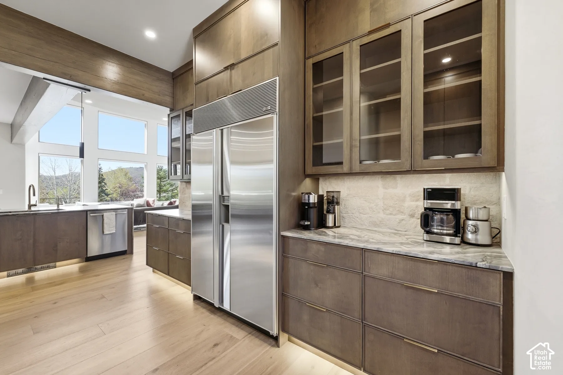 Integrated kitchen with wood-finish flooring, stainless steel appliances, dark wood cabinetry, light-toned stone countertops, and large picture windows
