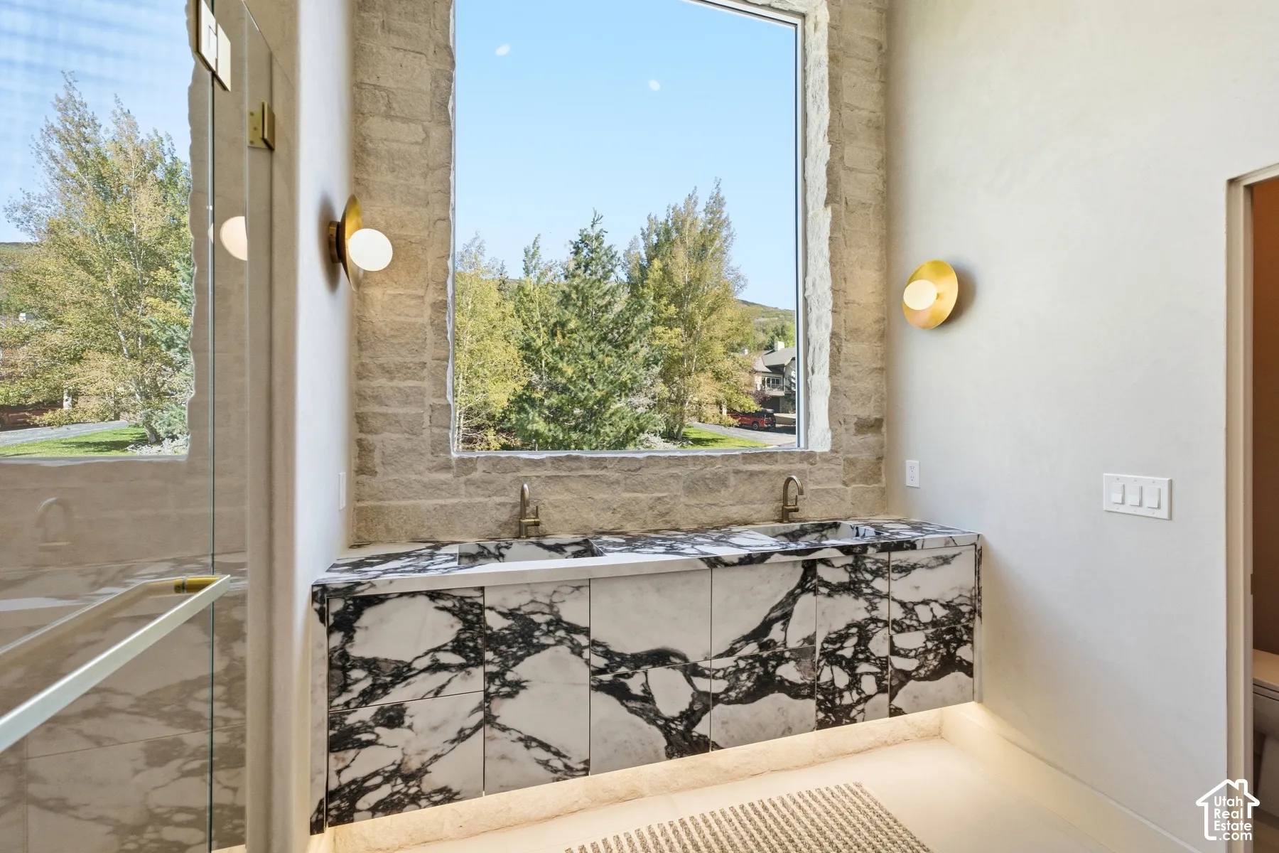 Stone accent wall with a large window, custom marble vanity, dual wall-mounted faucets, and contemporary wall sconces