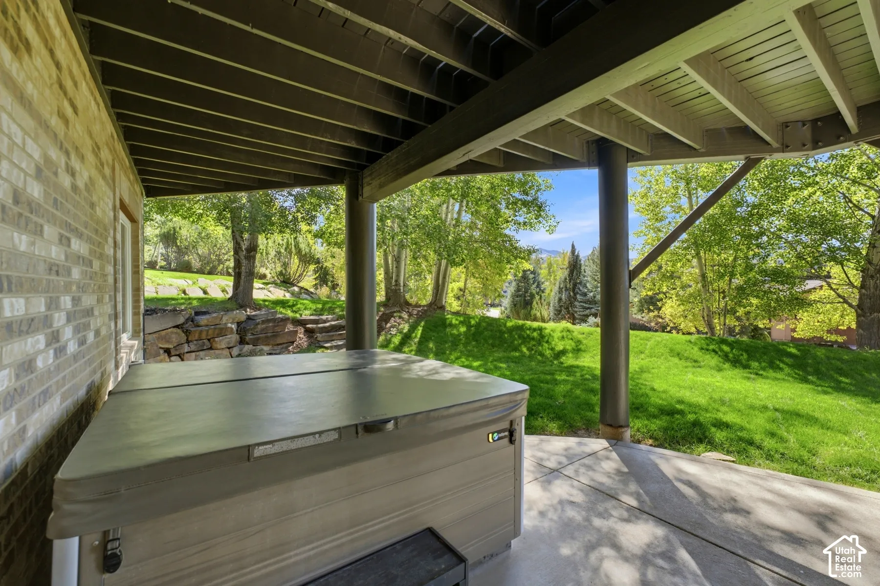 Covered patio with a built-in hot tub, concrete flooring, and a brick exterior wall, surrounded by mature trees and natural landscaping
