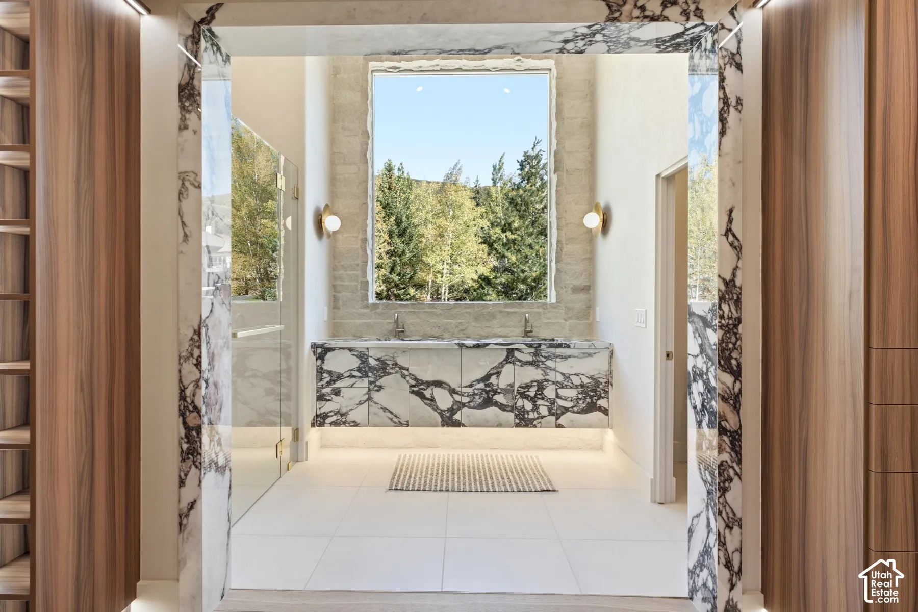 White tile flooring, custom wood-finish cabinetry, marble-clad archway, dual vanity with integrated sinks, and clear glass shower enclosure