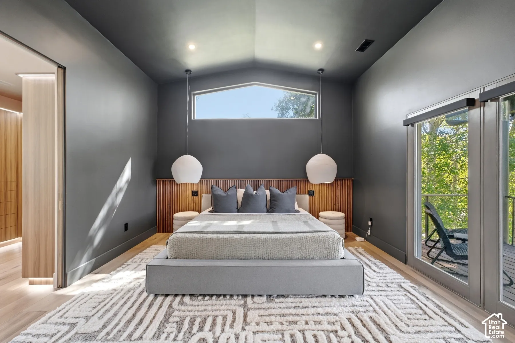 Vaulted ceiling with transom window, full-length glass doors, wood-finish flooring, built-in wood headboard, and pendant lighting