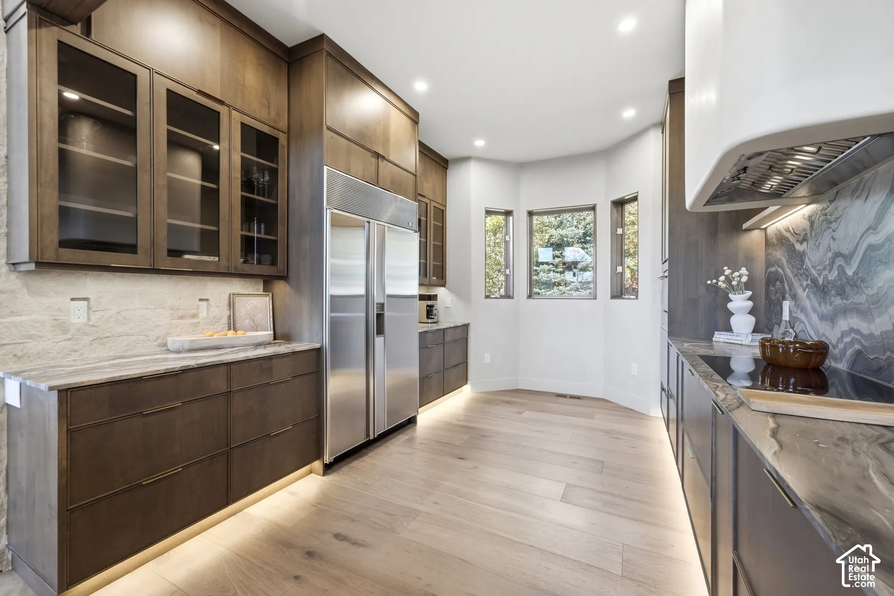 Wood-finish flooring with under-cabinet lighting, stainless steel refrigerator, built-in cooktop, and a marble backsplash