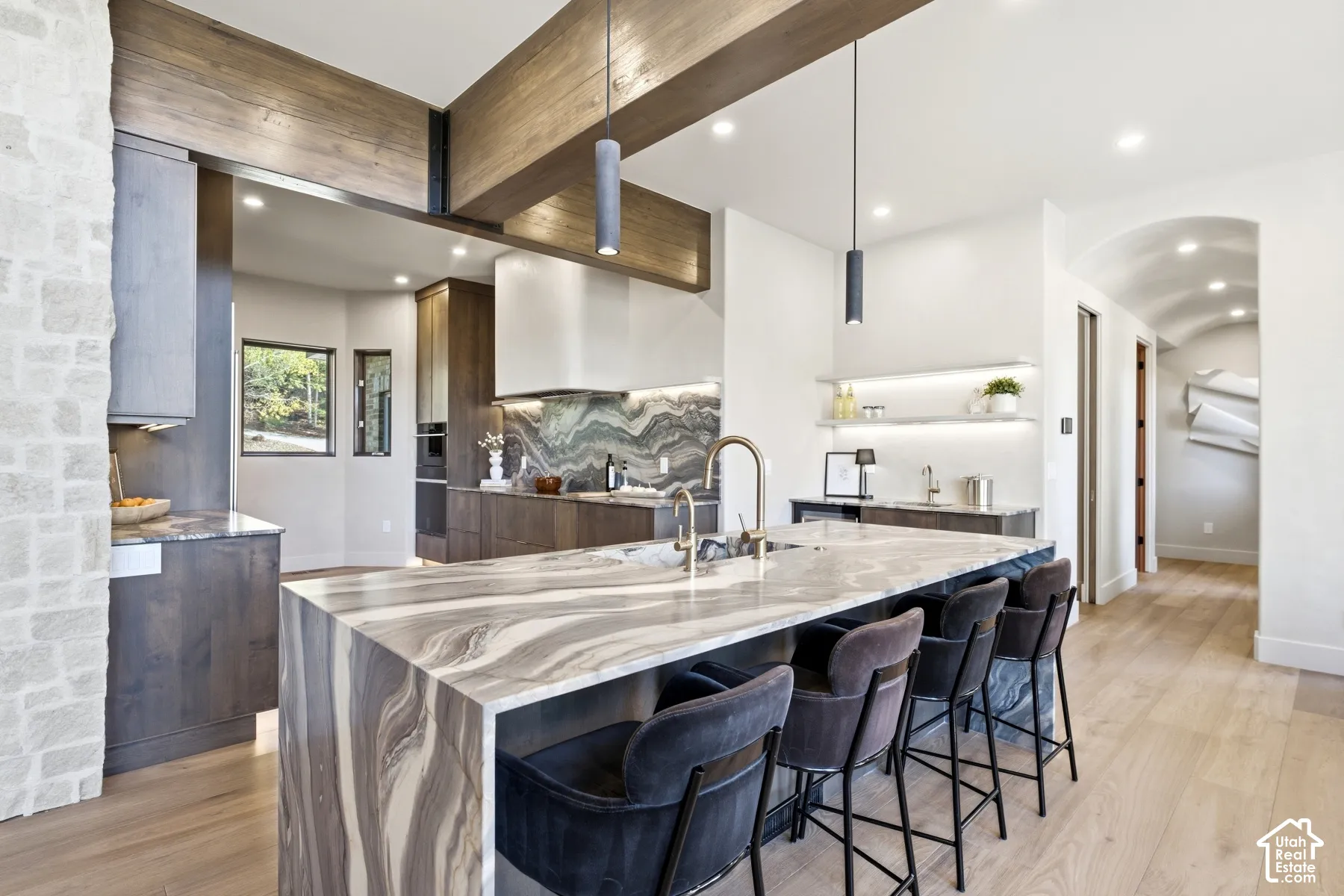 Gourmet kitchen featuring a large waterfall island with integrated seating, integrated range hood, wood-finish flooring, exposed wood beams, and a wet bar with floating shelves