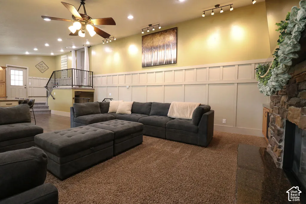 Stone-clad fireplace with dark hearth, carpeted flooring, track lighting, ceiling fan, and board and batten wainscoting