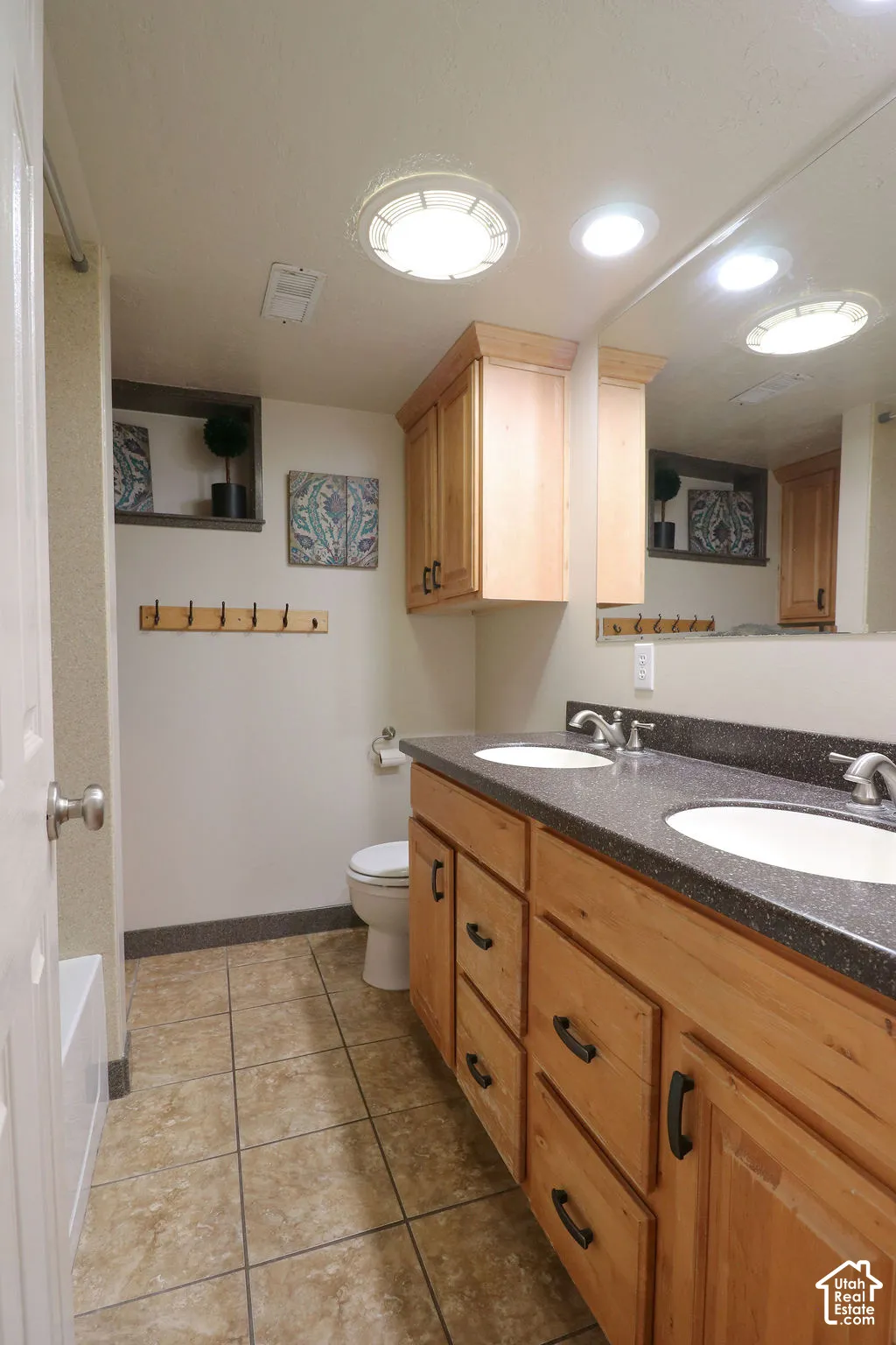 Dual sink vanity with wood-finish cabinetry, dark countertop, and tile flooring