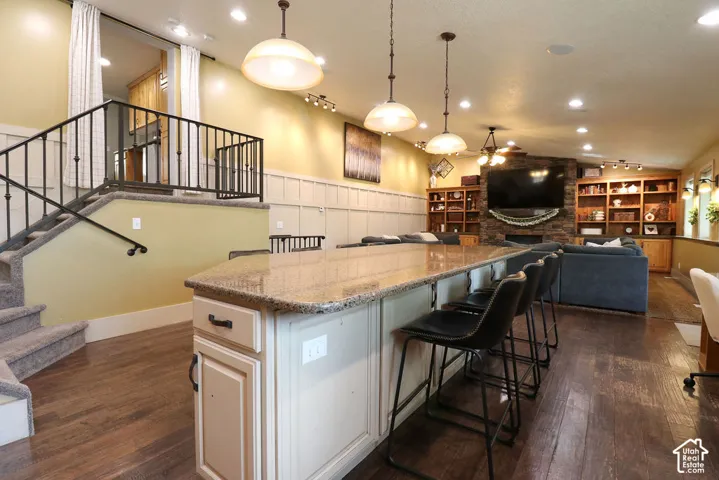 Granite-top island with pendant lighting, wood-finish flooring, built-in shelving, and a stone-clad fireplace
