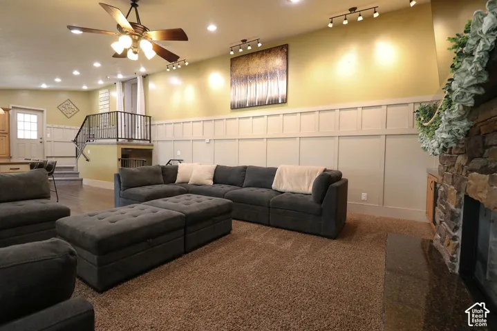 Stone-clad fireplace with dark hearth, carpeted flooring, track lighting, ceiling fan, and board and batten wainscoting