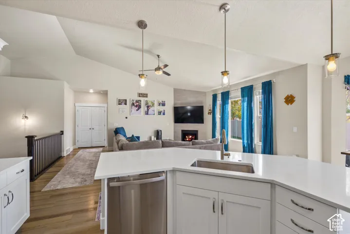 Vaulted ceilings with recessed lighting, kitchen island with integrated sink, stainless steel dishwasher, fireplace with stone surround, and wood-finish flooring