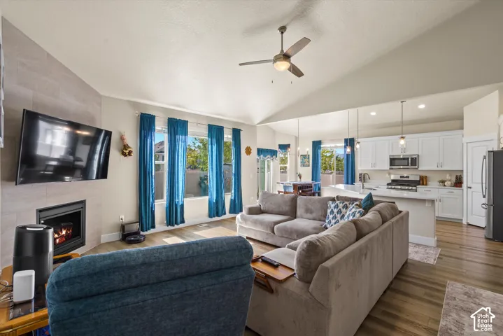 Vaulted ceiling with a ceiling fan, fireplace with a tile surround, kitchen island with pendant lighting, white cabinetry, and wood-finish flooring