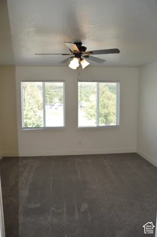 Textured ceiling, two windows with blinds, ceiling fan with integrated lighting, and dark grey carpet flooring