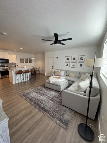 Open-concept living space featuring wood-finish flooring, a modern black ceiling fan, white cabinetry, stainless steel appliances, and a kitchen island with seating