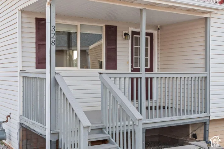 Covered porch with wood-finish railing and stairs, light-toned siding, and a dark-toned entry door with decorative glass panels