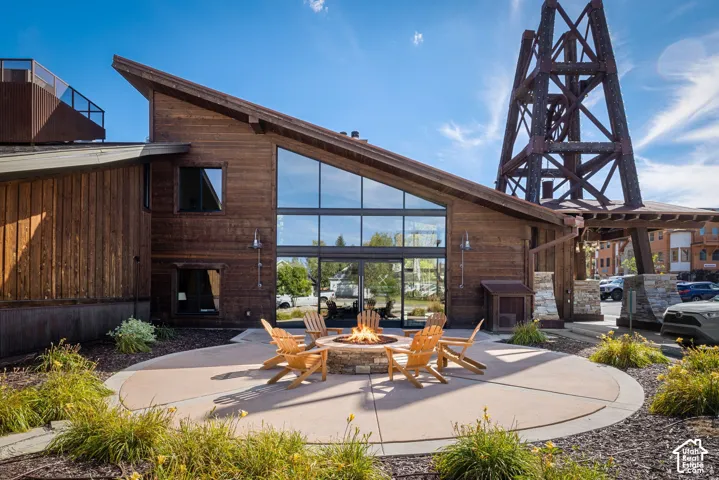 Wood-clad exterior with an angled roofline, extensive glazing, a circular concrete patio with an integrated fire pit, and a prominent industrial-style metal structure