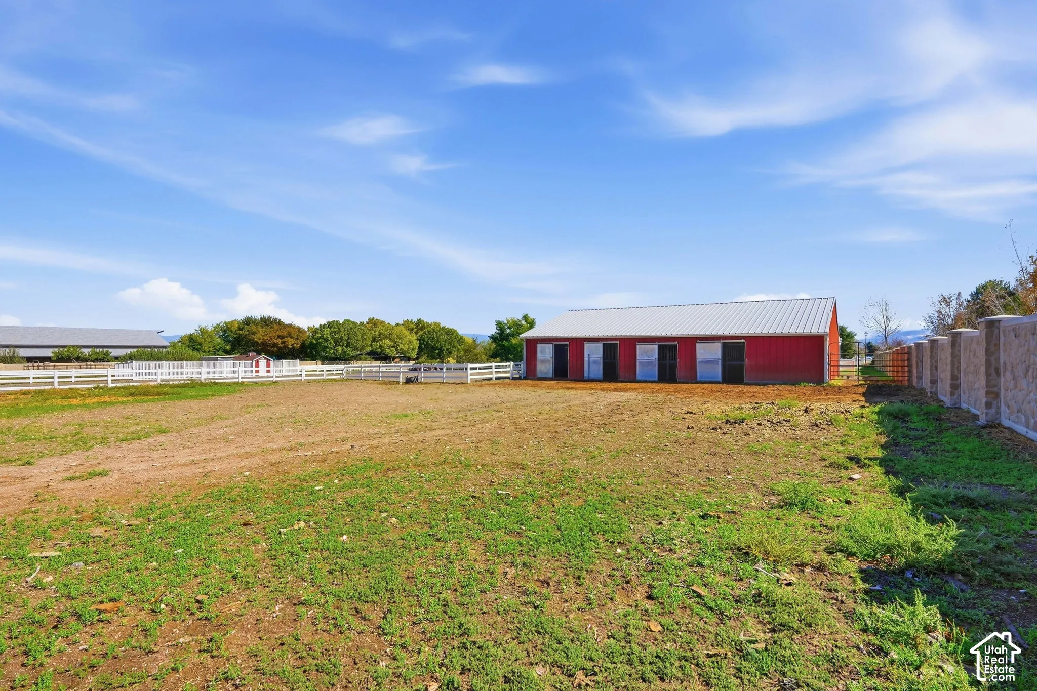 Red barn structure with a metal roof, multiple sliding doors, white fencing, and a concrete-block privacy wall