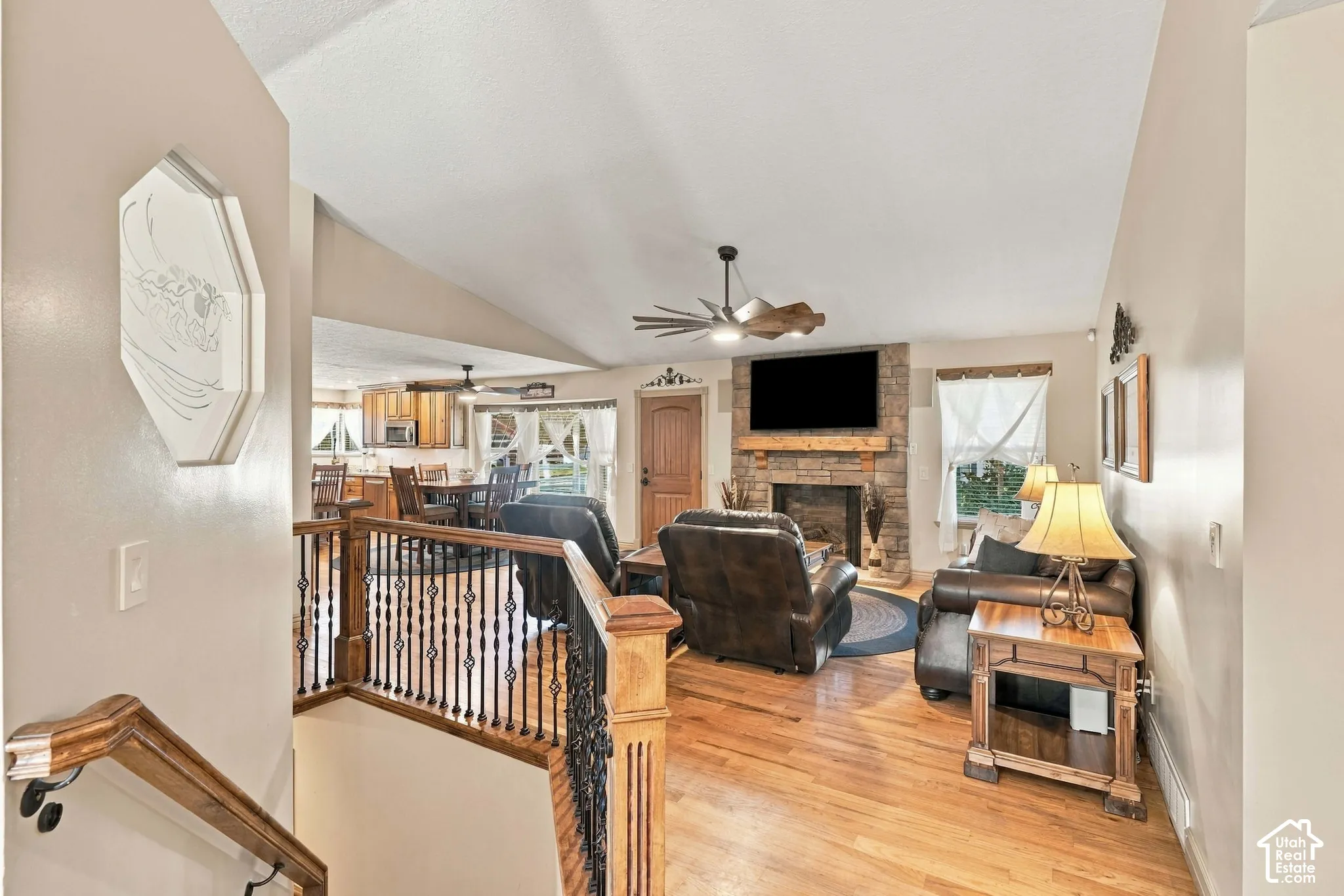 Vaulted ceilings with a ceiling fan, a stone-clad fireplace with a wood mantel, wood-finish flooring, and a wood and wrought iron staircase railing