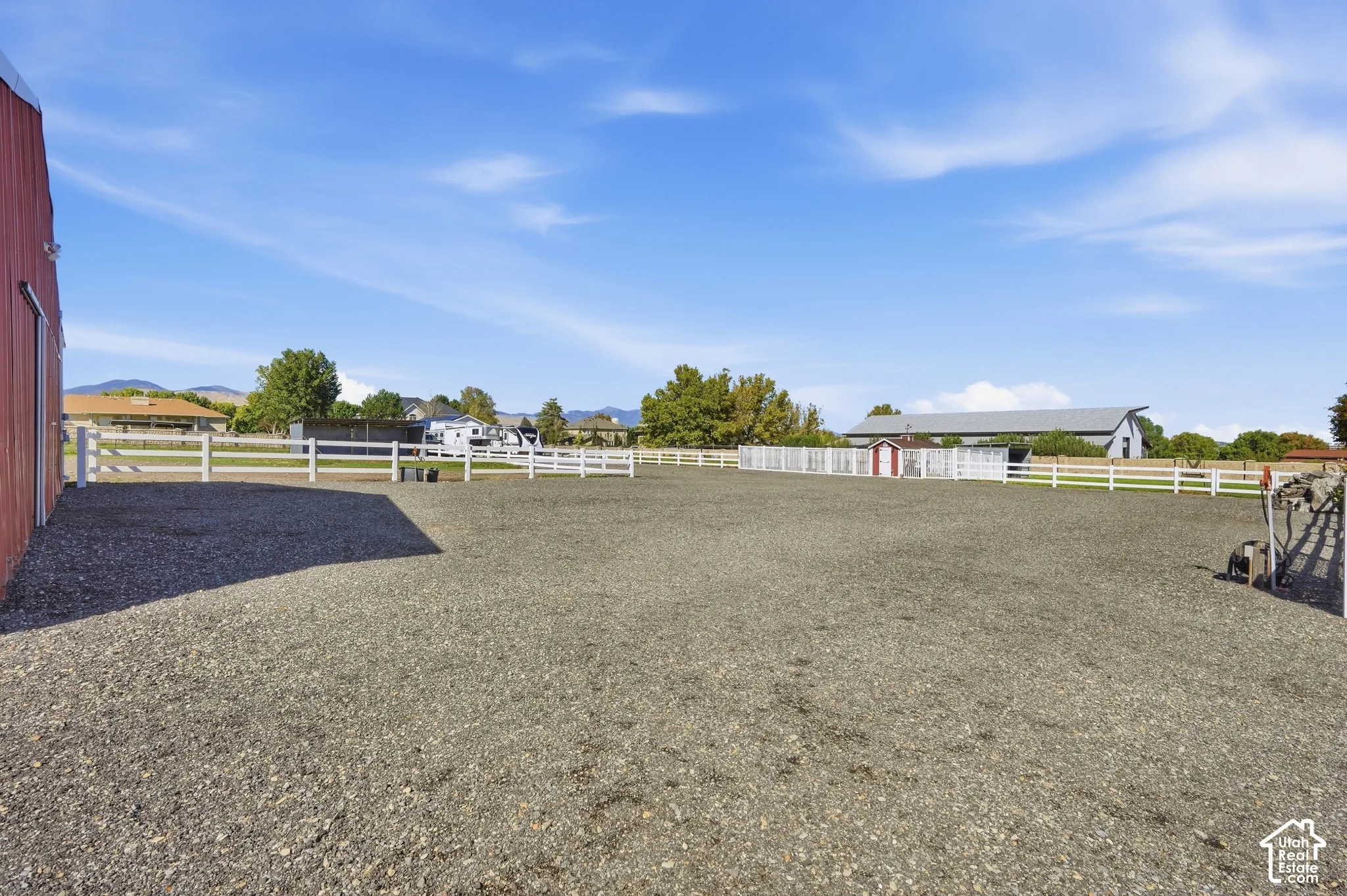 Expansive gravel-surfaced equestrian arena featuring white fencing, with a red barn structure and additional outbuildings visible