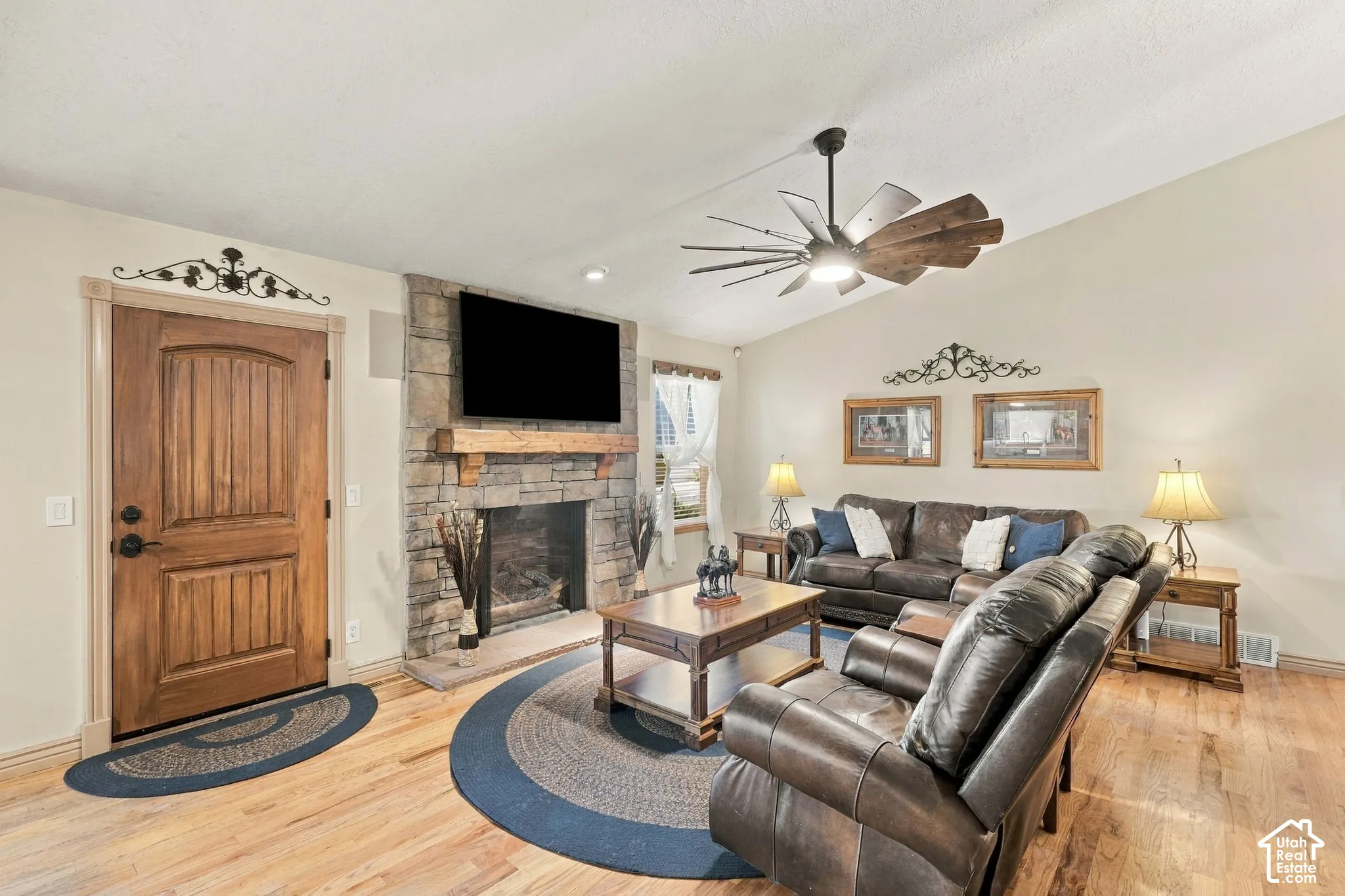 Vaulted ceiling, stone-clad fireplace with wood mantle, wood-finish flooring, wood-panel entry door, and a ceiling fan with integrated lighting