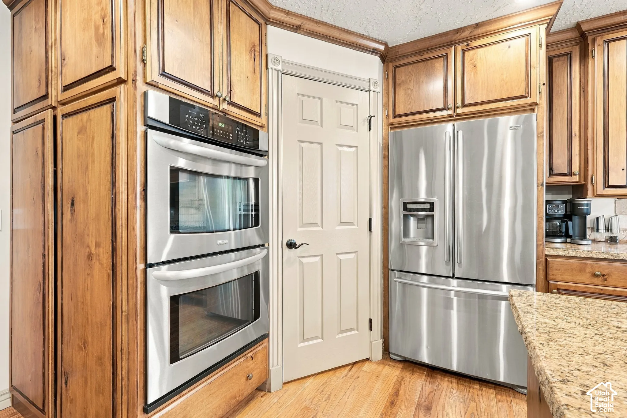 Wood cabinetry with crown molding, stainless steel double wall ovens, stainless steel French door refrigerator, light-toned wood-finish flooring, and granite-style countertops