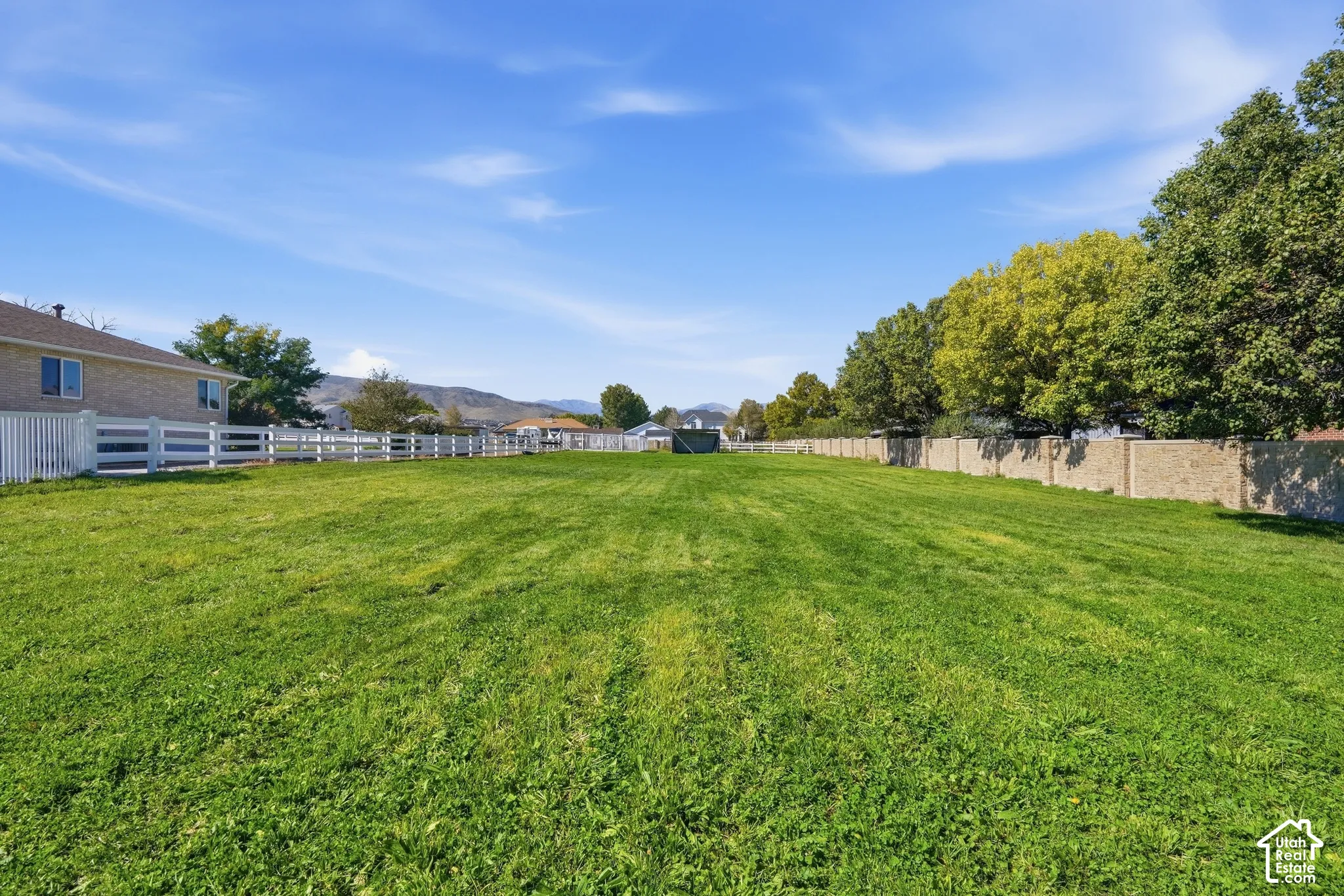 Expansive green space featuring a white multi-rail fence, a stone-finish privacy wall, and mature perimeter trees