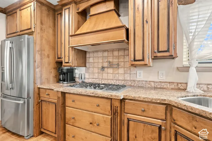 Full-height wood cabinetry with crown molding, integrated range hood, stainless steel refrigerator, tile backsplash, and composite countertops