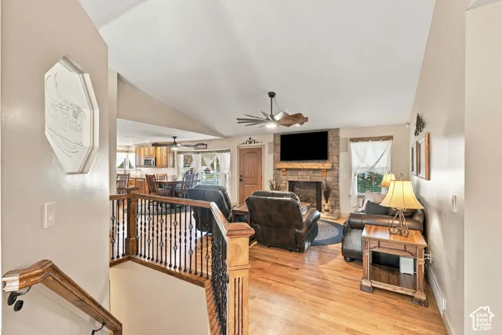 Vaulted ceilings with a ceiling fan, a stone-clad fireplace with a wood mantel, wood-finish flooring, and a wood and wrought iron staircase railing