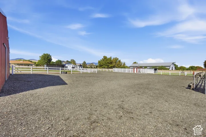 Expansive gravel-surfaced equestrian arena featuring white fencing, with a red barn structure and additional outbuildings visible