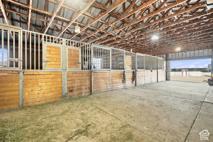Barn interior featuring multiple stalls with wood and metal construction, exposed wooden rafters, corrugated metal roofing, and overhead lighting fixtures
