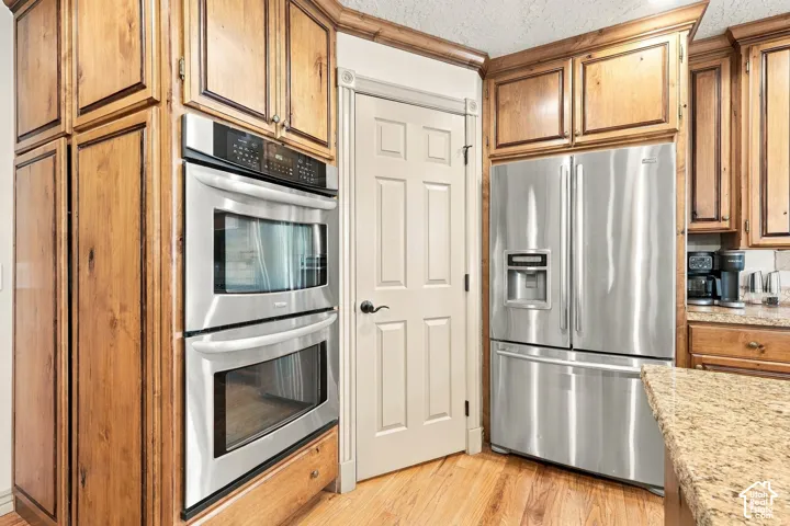 Wood cabinetry with crown molding, stainless steel double wall ovens, stainless steel French door refrigerator, light-toned wood-finish flooring, and granite-style countertops