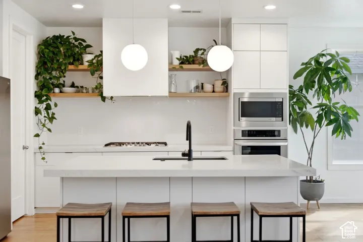 White kitchen featuring a gas range, white cabinetry, integrated stainless steel appliances, a center island with sink, and white pendant lighting