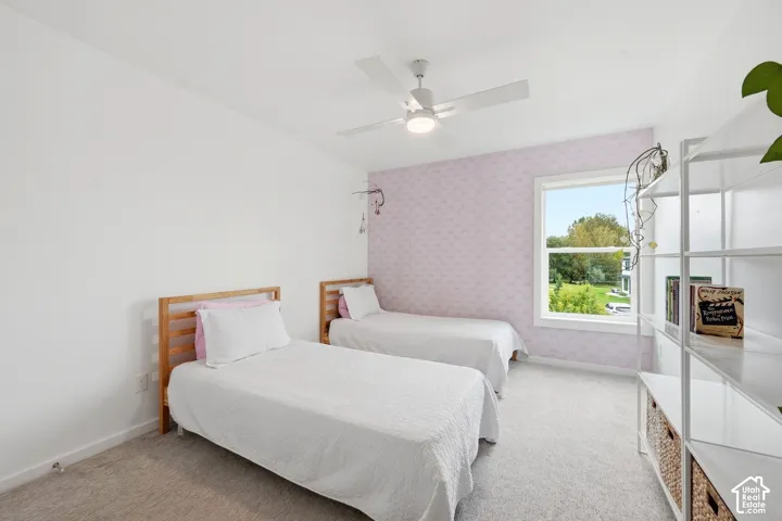 Window with exterior scenery, overhead ceiling fan, white walls, patterned accent wall, and light-colored carpeting