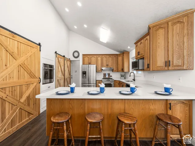 Vaulted ceiling with recessed lighting, wood-finish flooring, wood cabinetry, stainless steel appliances, and a kitchen island with a light-toned countertop