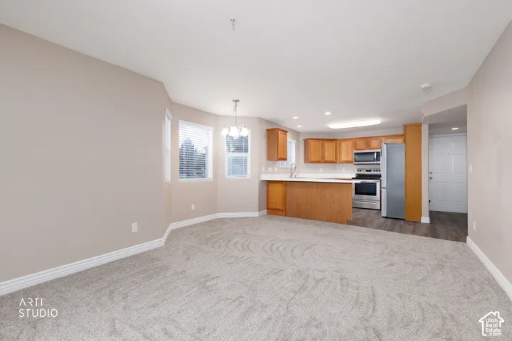 Open-concept living space featuring light-toned walls, plush carpeting, and a kitchen with wood-finish cabinetry, stainless steel appliances, and a peninsula countertop