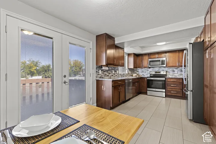Kitchen featuring wood cabinetry, stainless steel appliances, mosaic tile backsplash, light-toned tile flooring, and French doors leading to an outdoor deck