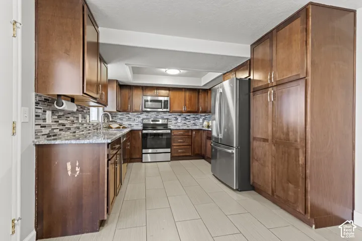 Kitchen featuring wood-finish cabinetry, tile backsplash, stainless steel appliances, and light-toned floor tiling