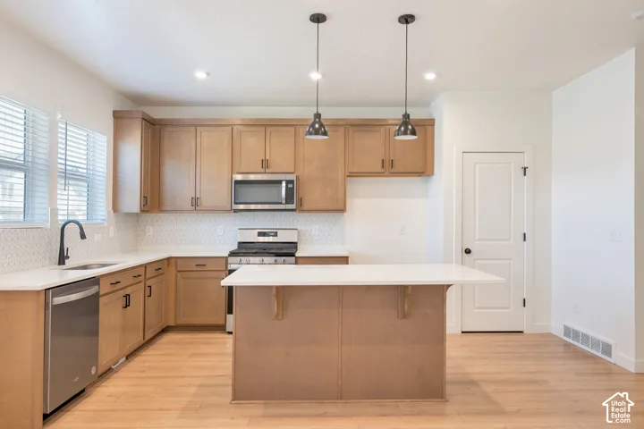 Kitchen featuring light wood-finish cabinetry, white countertops, a mosaic tile backsplash, stainless steel appliances, and a central island with pendant lighting