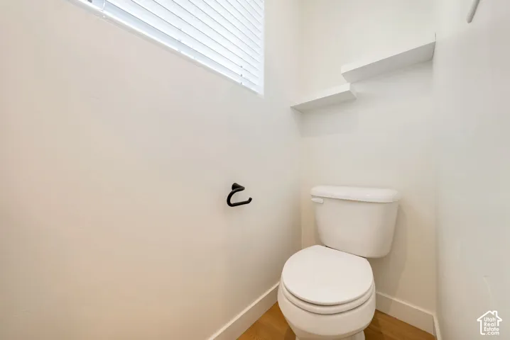 White toilet with two floating shelves, wood-finish flooring, and a window with blinds