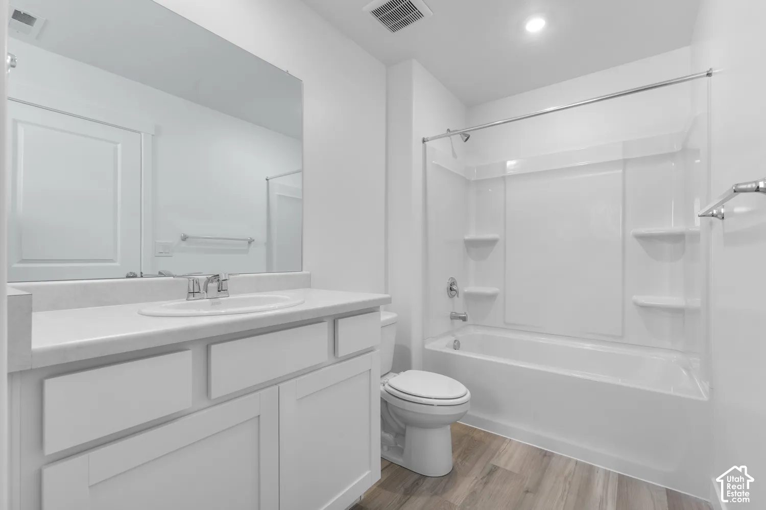 White vanity with a white countertop and integrated sink, a large wall-mounted mirror, a built-in bathtub with a shower and integrated shelving, and wood-finish flooring