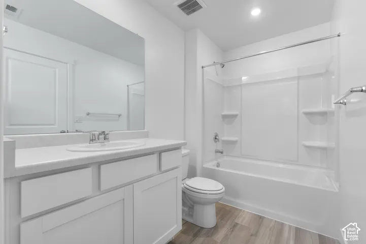 White vanity with a white countertop and integrated sink, a large wall-mounted mirror, a built-in bathtub with a shower and integrated shelving, and wood-finish flooring