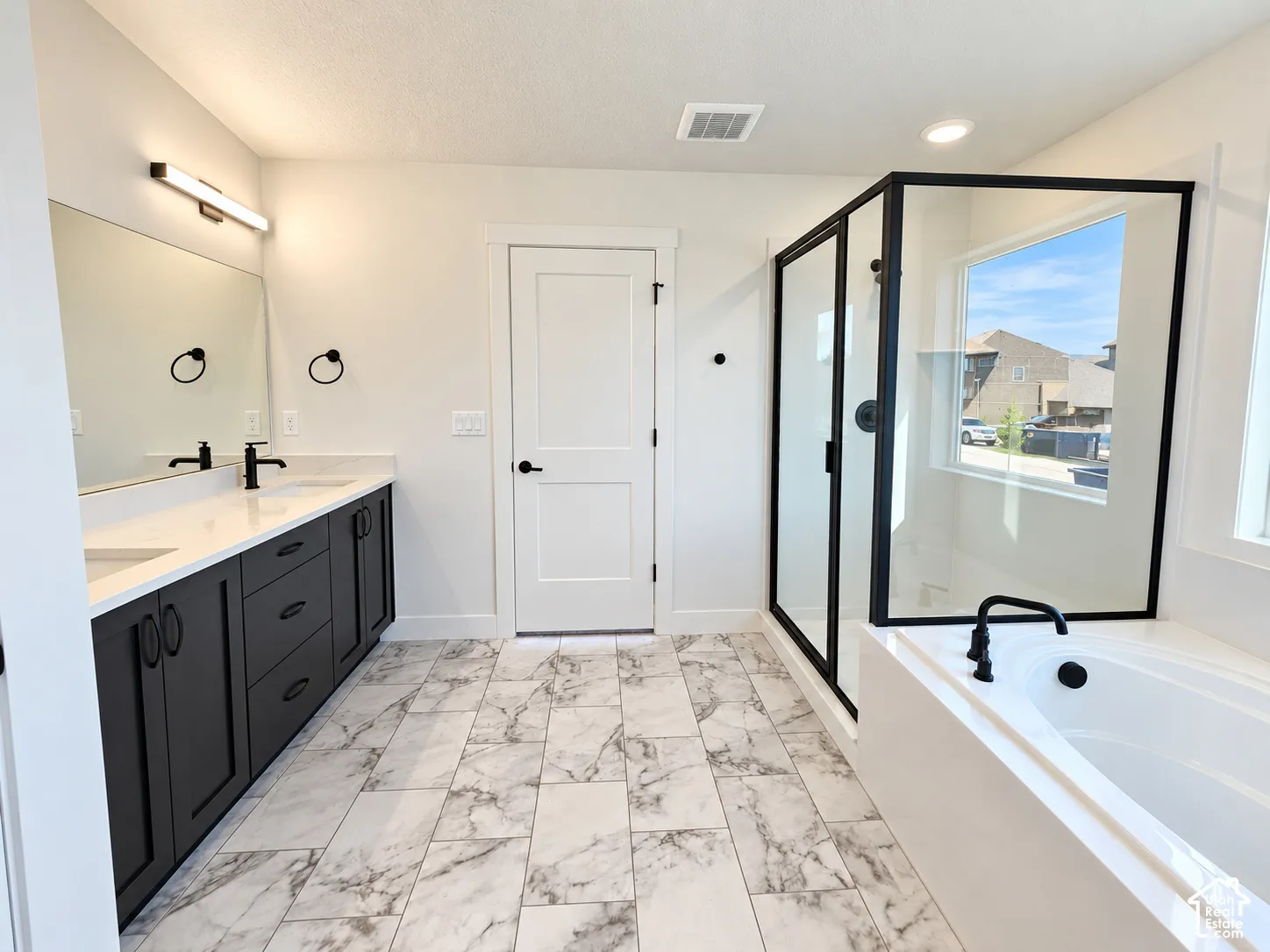 Spacious bathroom featuring a dual vanity with dark cabinetry, a built-in soaking tub, a separate glass-enclosed shower, and light-toned tile flooring