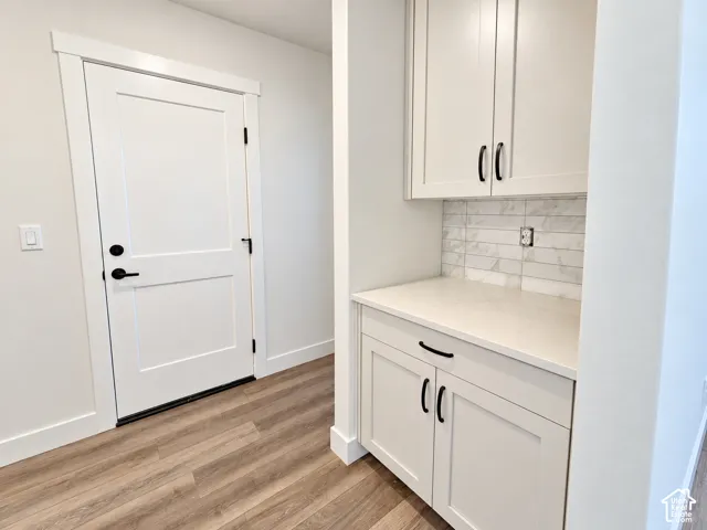 White panel door, wood-finish flooring, white cabinetry with black hardware, light-toned countertop, and tile backsplash