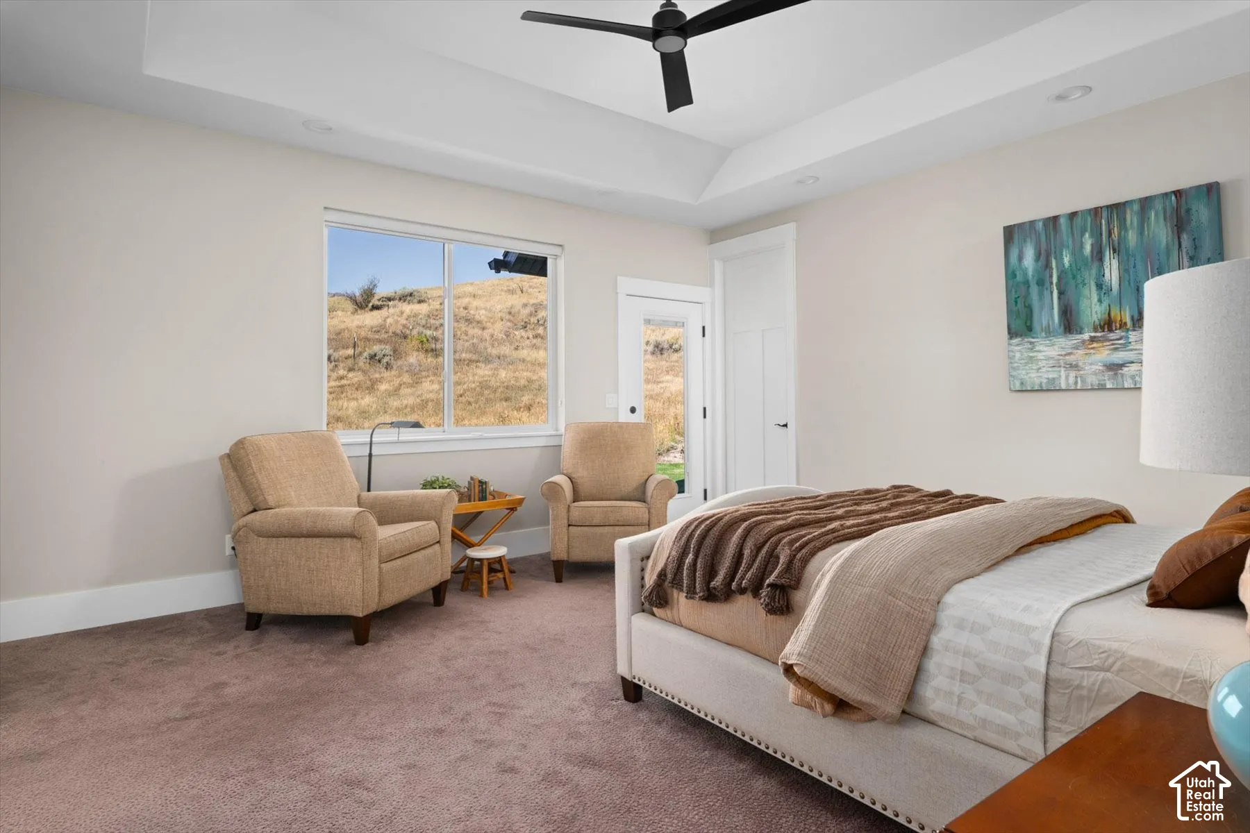 Spacious primary bedroom featuring expansive windows with natural light, carpeted flooring, a ceiling fan, and a dedicated sitting area with upholstered seating