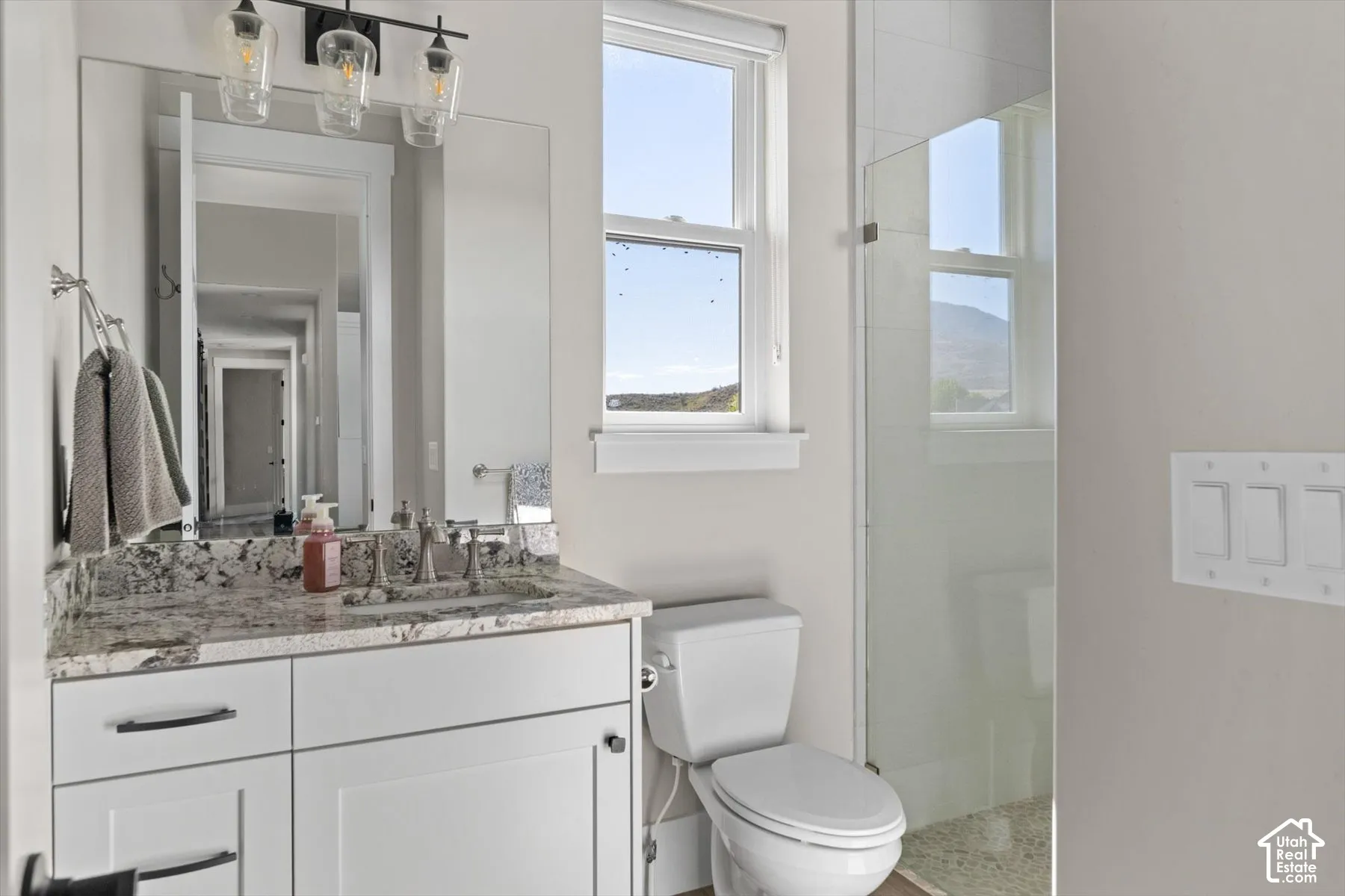 Bathroom featuring a vanity with a stone countertop, an undermount sink, a glass-enclosed shower, a toilet, and a window