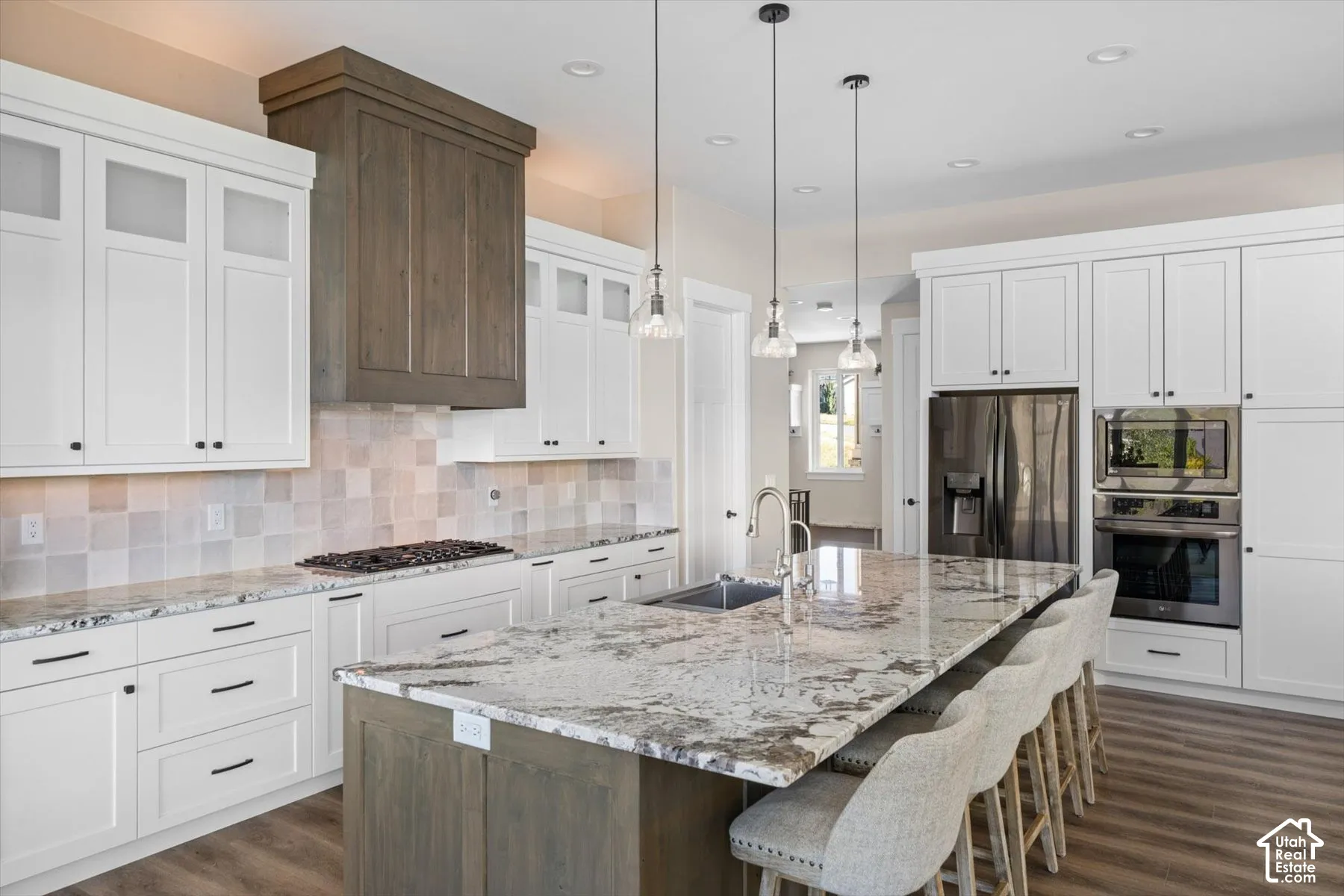 Kitchen featuring white shaker-style cabinetry, a wood-finish range hood, tile backsplash, and a multi-level island with a built-in sink and pendant lighting